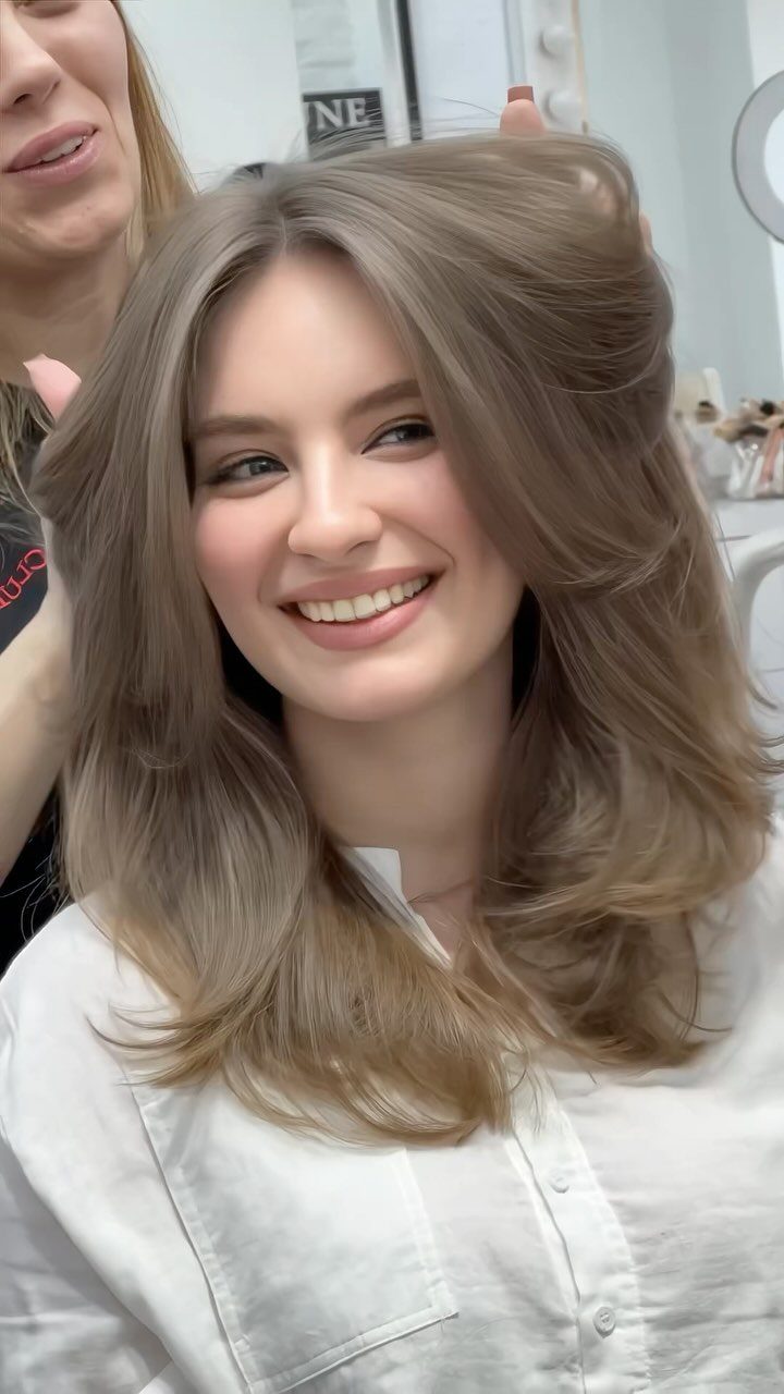 7 Clean Look Hair Ideas for Sleek Buns and Ponytails 5 Woman with voluminous light brown hair and loose face-framing strands smiling in a salon.