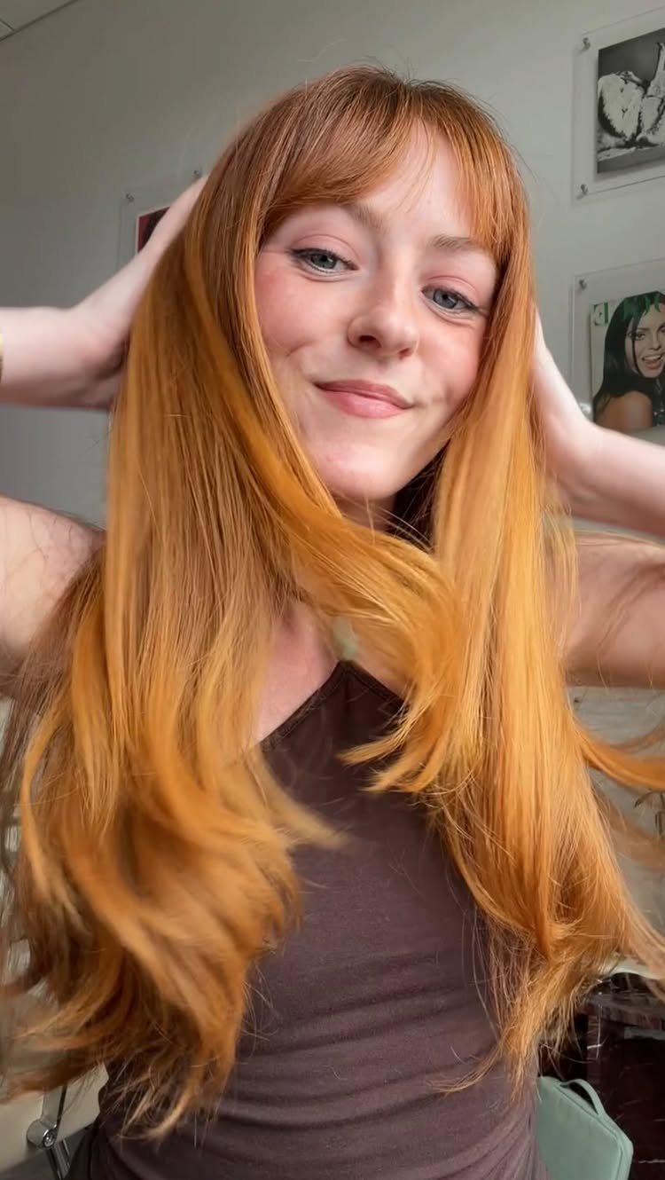 Cowboy Hair Color Ideas: 6 Copper Shades to Try 1 Woman with long wavy copper hair and bangs posing in a brown top.