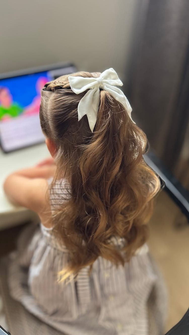 17 Cute Hairstyles for Picture Day, From Braids to Buns 15 Young girl with wavy brown hair wearing a white bow and small braid.