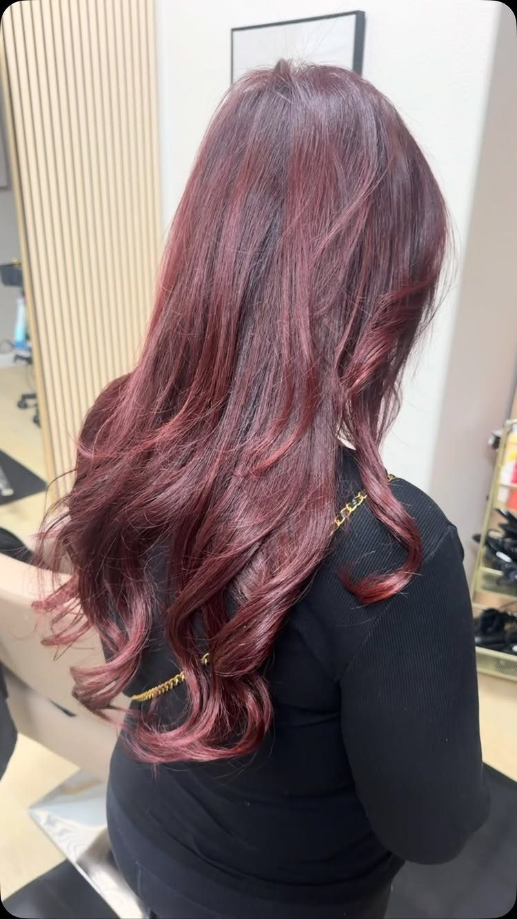 Woman with long wavy dark maroon hair seen from behind in a salon.