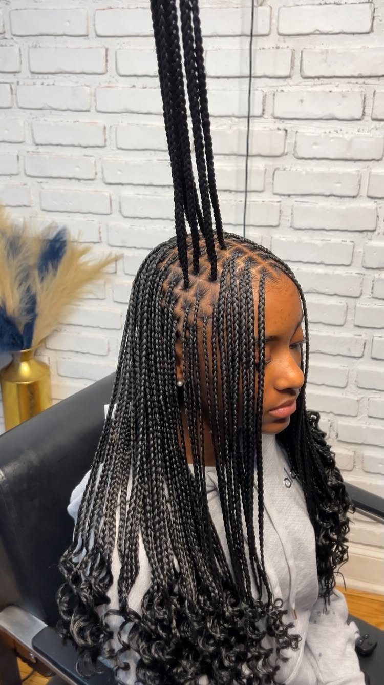 Woman with long black feed-in braids and curly ends against white brick wall