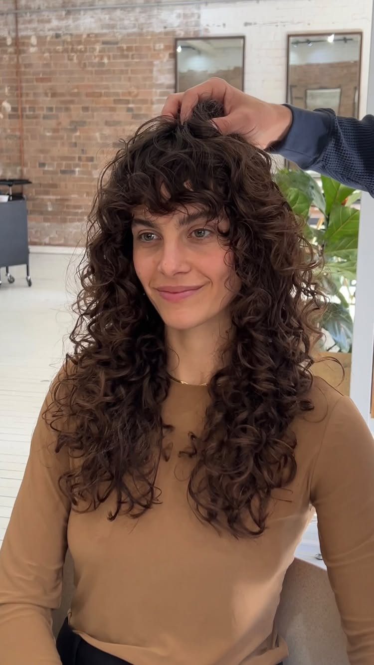 12 Good Hairstyles for Curly Hair, From Bangs to Braids 1 Woman with long brown curly hair and curly bangs in a salon setting