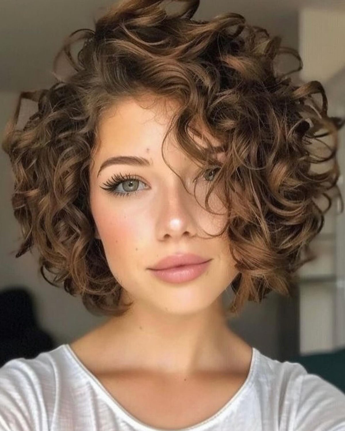 12 Good Hairstyles for Curly Hair, From Bangs to Braids 6 Woman with a short, voluminous brown curly bob hairstyle and a white shirt.