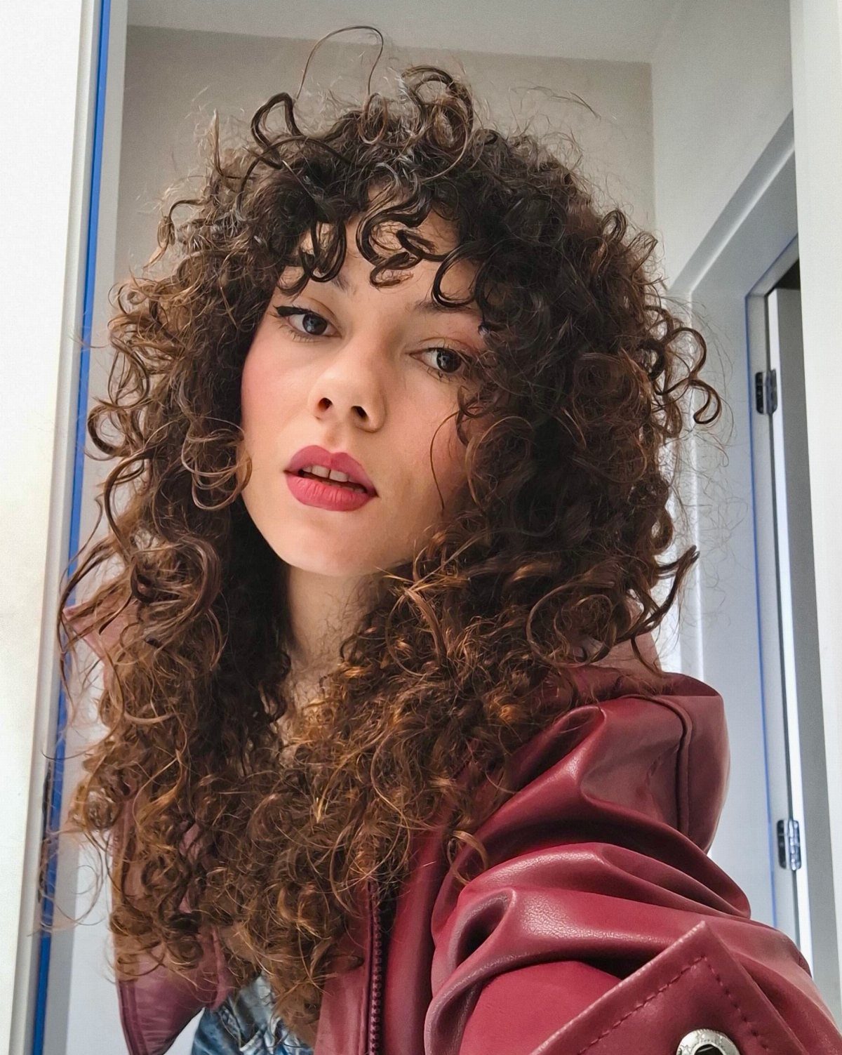 12 Good Hairstyles for Curly Hair, From Bangs to Braids 2 Woman with a curly shag haircut and bangs wearing a red leather jacket.