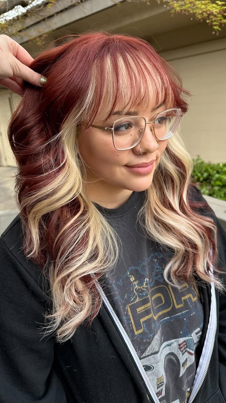22 Hair Color Red and Blonde Ideas for Every Length 8 Woman with deep red wavy hair and thick blonde face-framing highlights wearing glasses.
