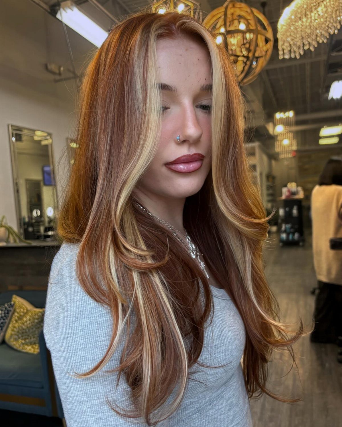 22 Hair Color Red and Blonde Ideas for Every Length 5 Woman with long copper-red hair and bright blonde face-framing highlights in a salon.