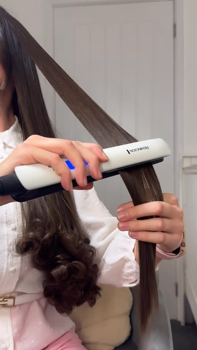 Person using a white flat iron to straighten long brown hair at home