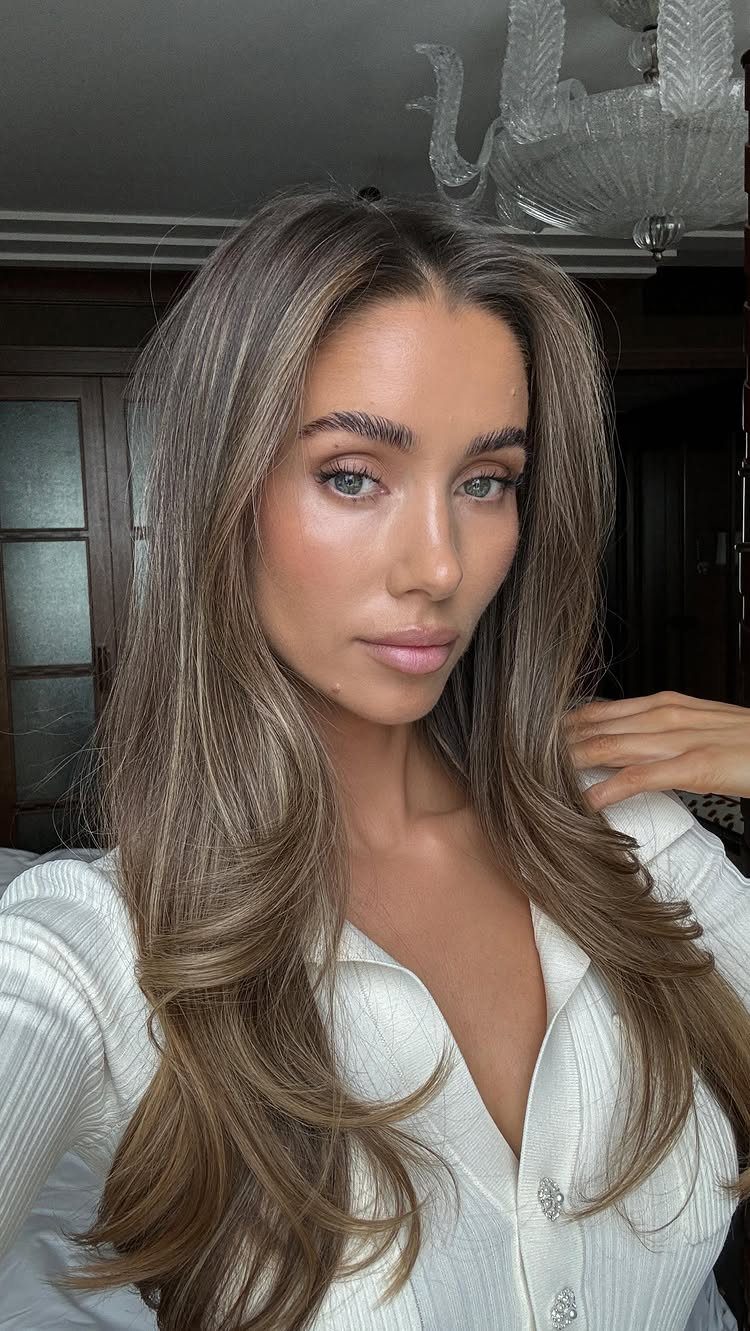 Woman with long styled ash-brown hair wearing a white ribbed top indoors