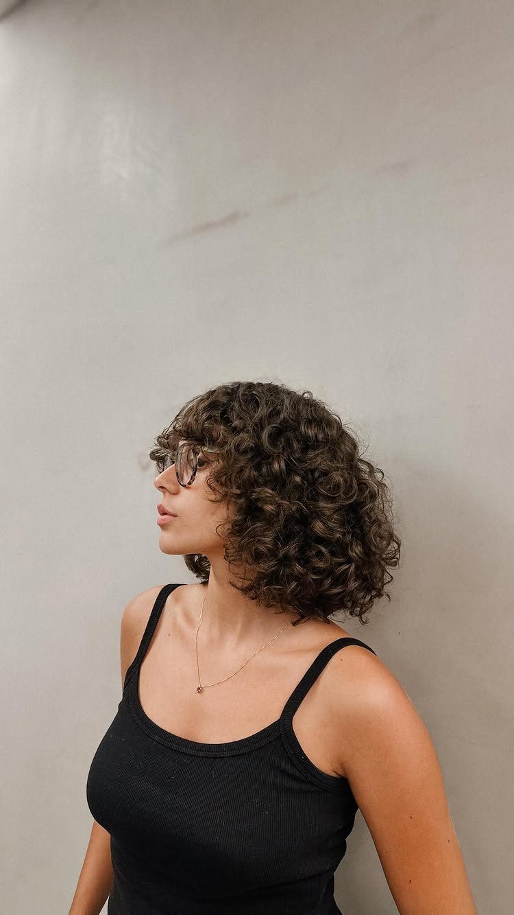 Woman with a curly brown bob haircut and glasses against a grey wall