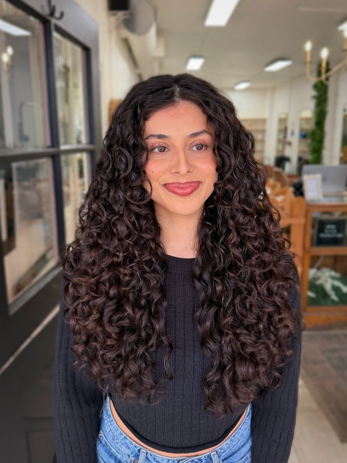 Woman with long, voluminous dark curly hair wearing a black top and jeans.
