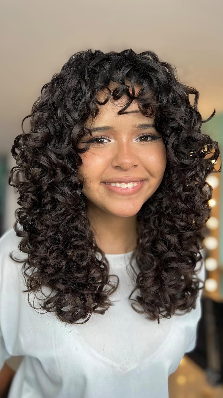 Woman with voluminous dark brown curly hair and bangs smiling in a white shirt.