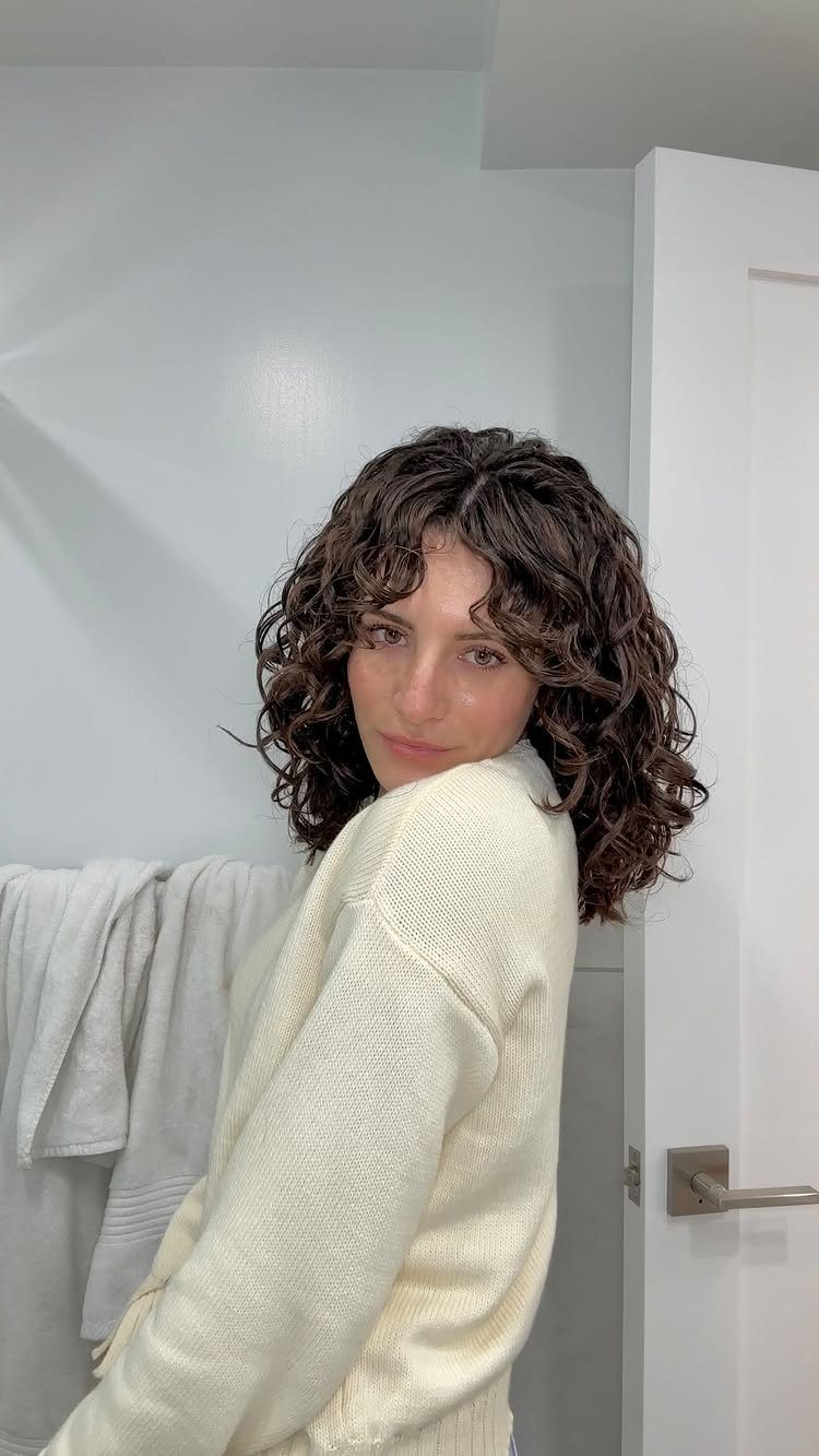 Woman with shoulder-length dark brown curly hair wearing a cream knit sweater.