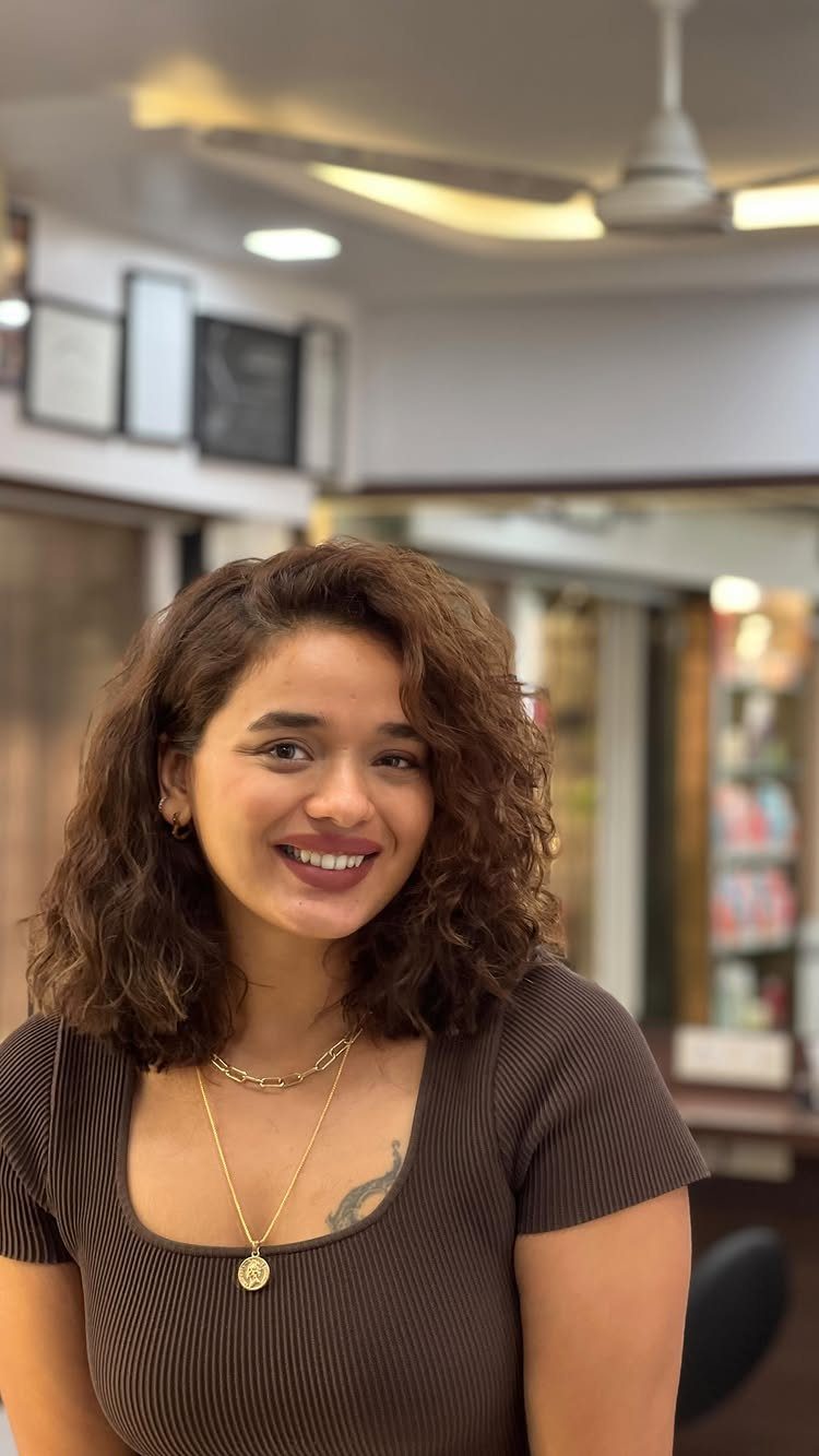 Woman with a textured lob haircut featuring curly brown hair and a smile.