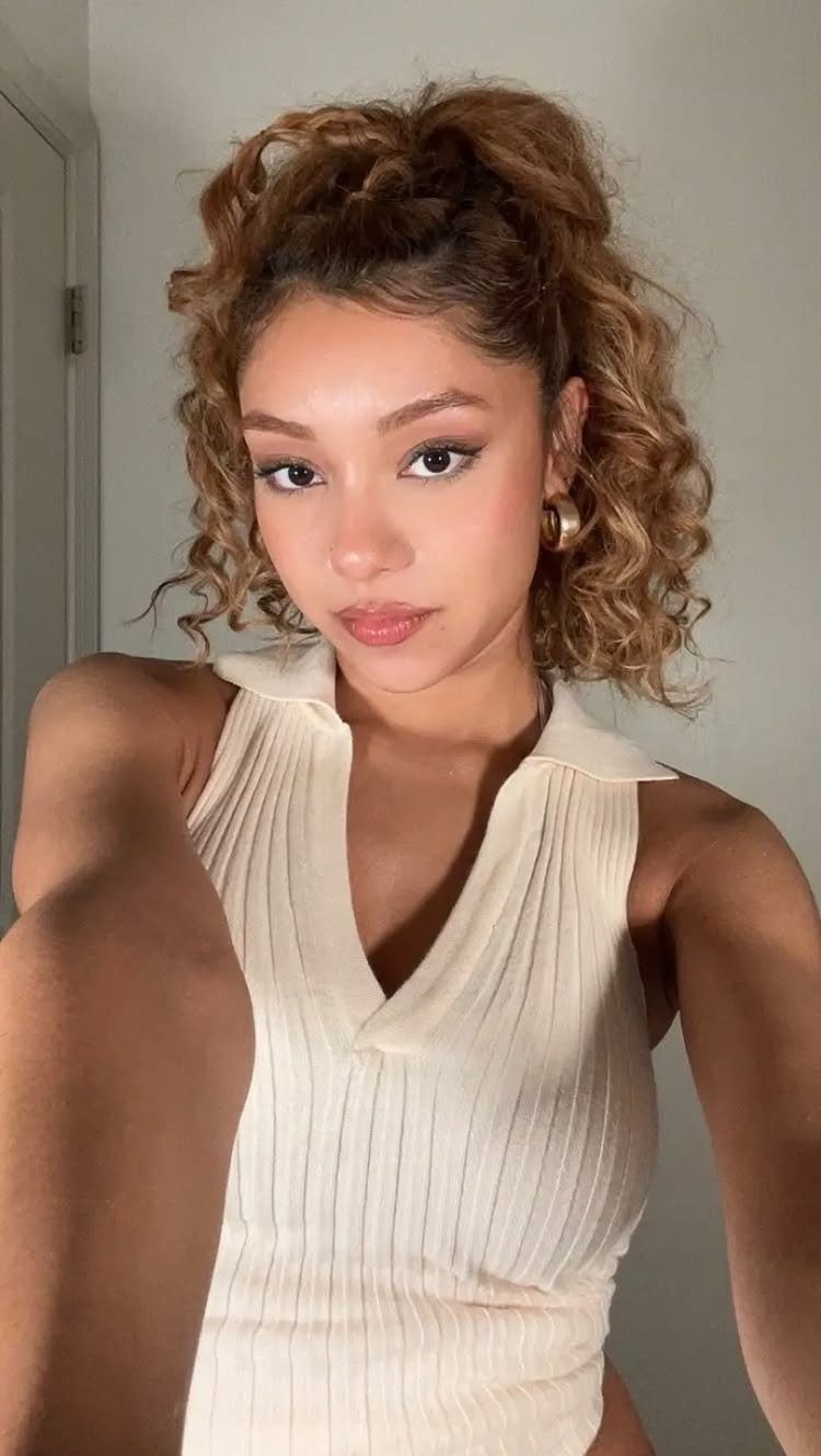 Woman with light brown curly hair in a high ponytail and white top.