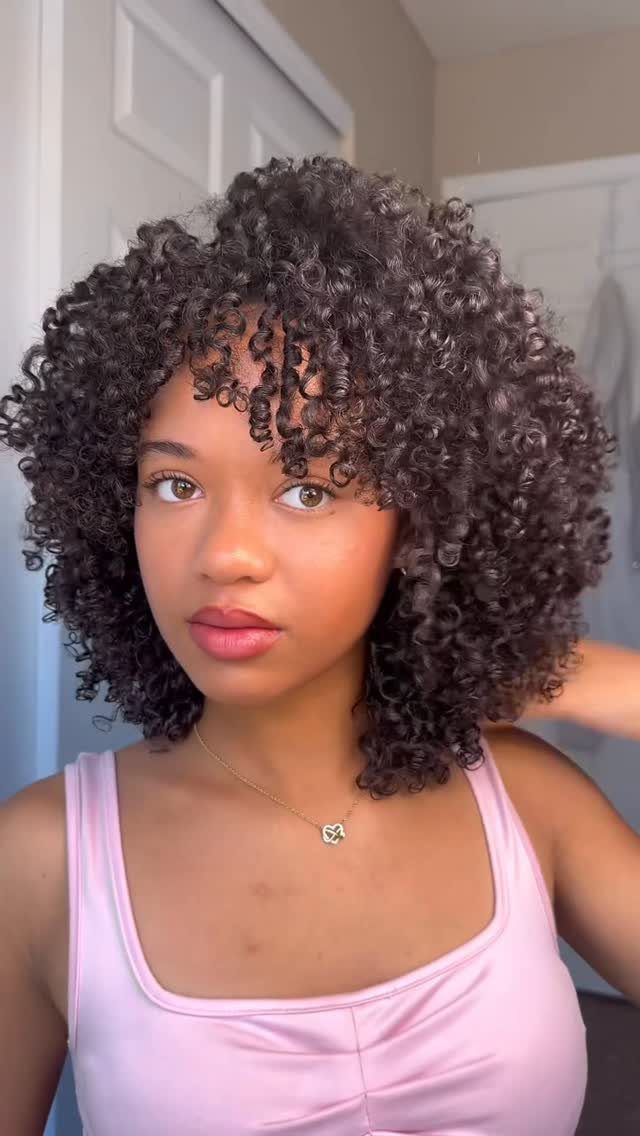 Woman with voluminous dark curly hair and bangs wearing a pink satin top.