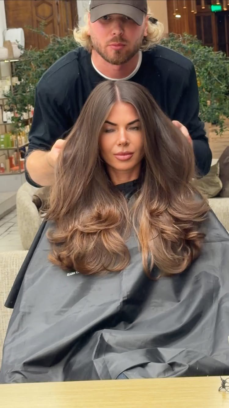 20 Long Hair Hairstyles for Work, From Buns to Braids 6 Woman with voluminous brown blowout curls being held by a stylist in a salon