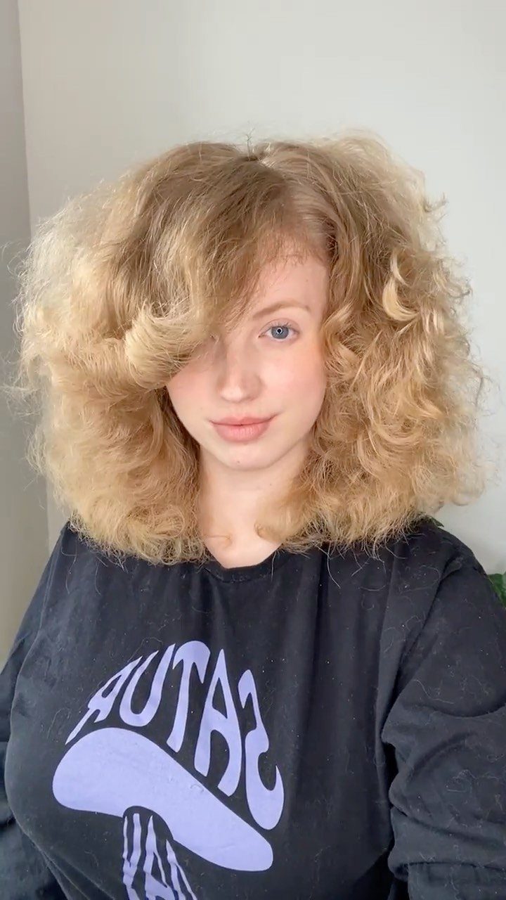 8 Loose Curl Hair Ideas for Blonde, Auburn and Long Looks 2 Woman with voluminous blonde brushed-out curls and bangs wearing a black graphic t-shirt.