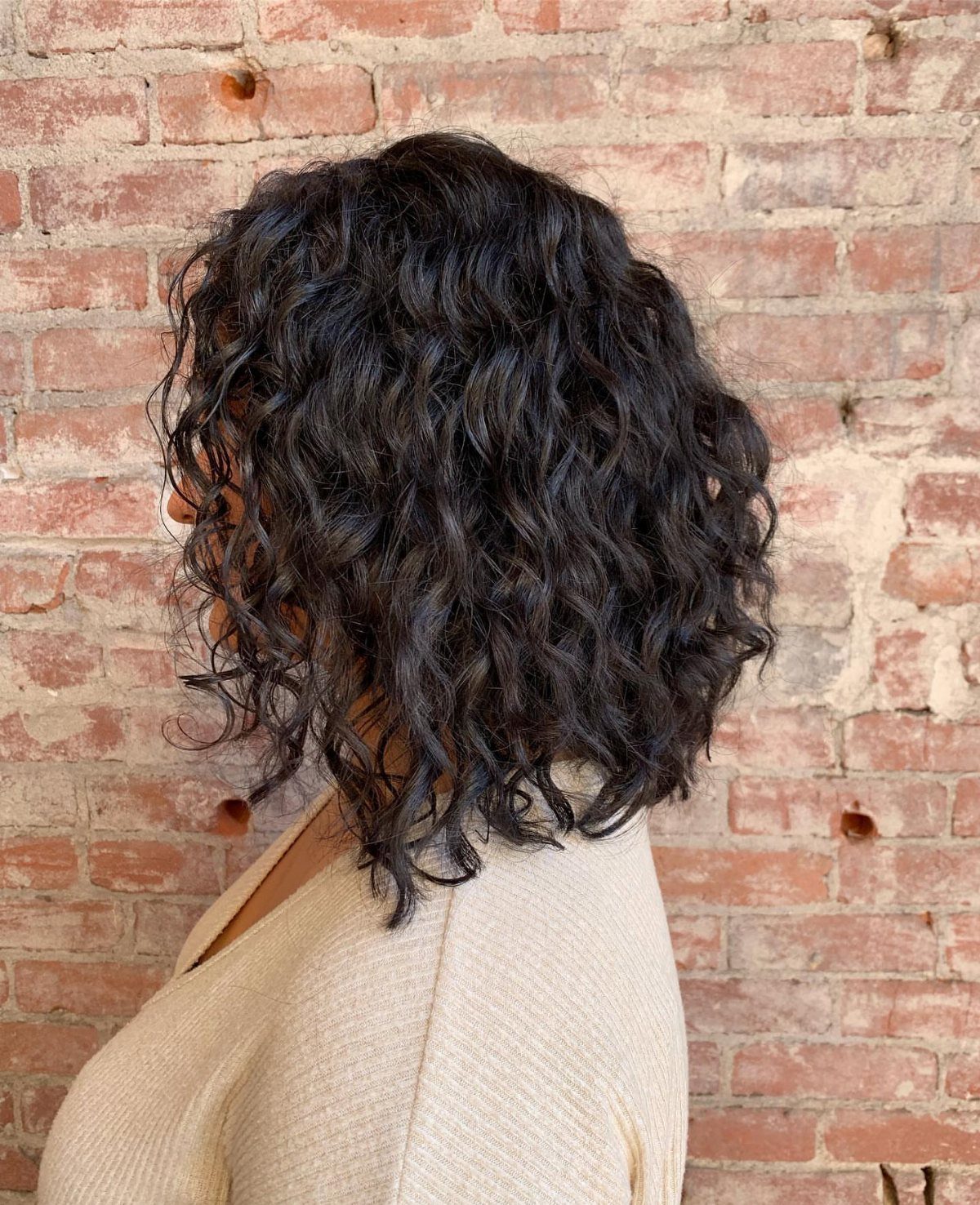 11 Medium Length Curly Hair Hairstyles for Every Look 4 Dark curly shoulder-length hair styled in a lob against a red brick wall