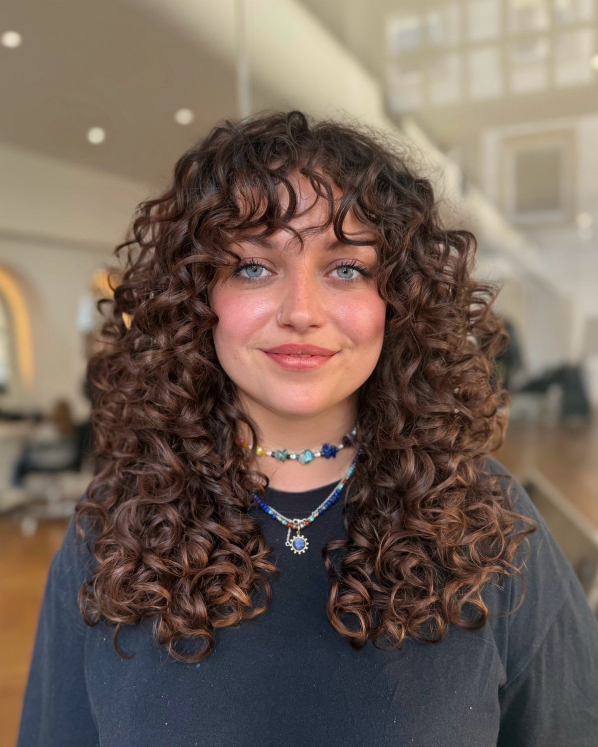 11 Medium Length Curly Hair Hairstyles for Every Look 7 Woman with voluminous medium-length brown curly hair and face-framing ringlets and bangs.