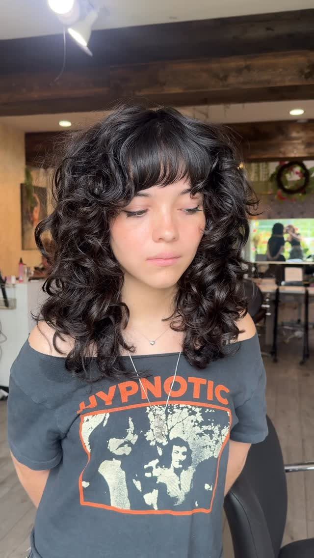 11 Medium Length Curly Hair Hairstyles for Every Look 5 Woman with voluminous medium-length dark curly hair and full bangs in a salon.