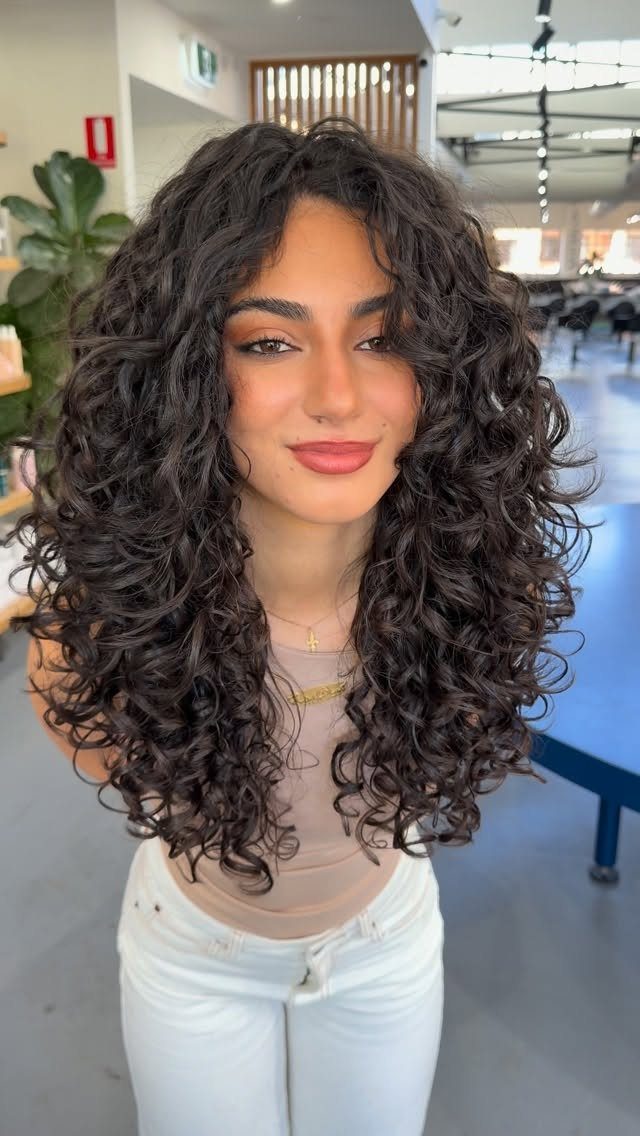 11 Medium Length Curly Hair Hairstyles for Every Look 6 Woman with voluminous dark brown curly hair in a layered mid-length hairstyle.