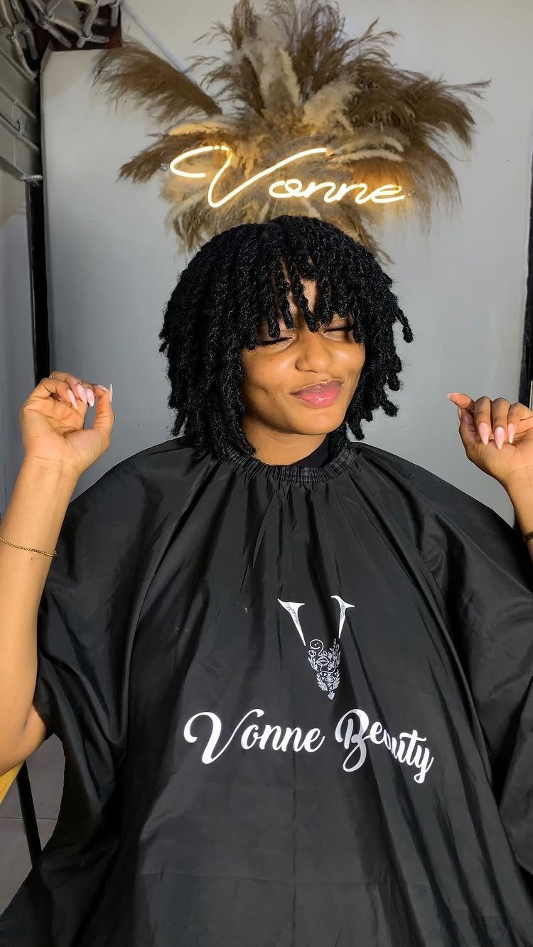 Woman with short black mini twists and bangs wearing a black salon cape.