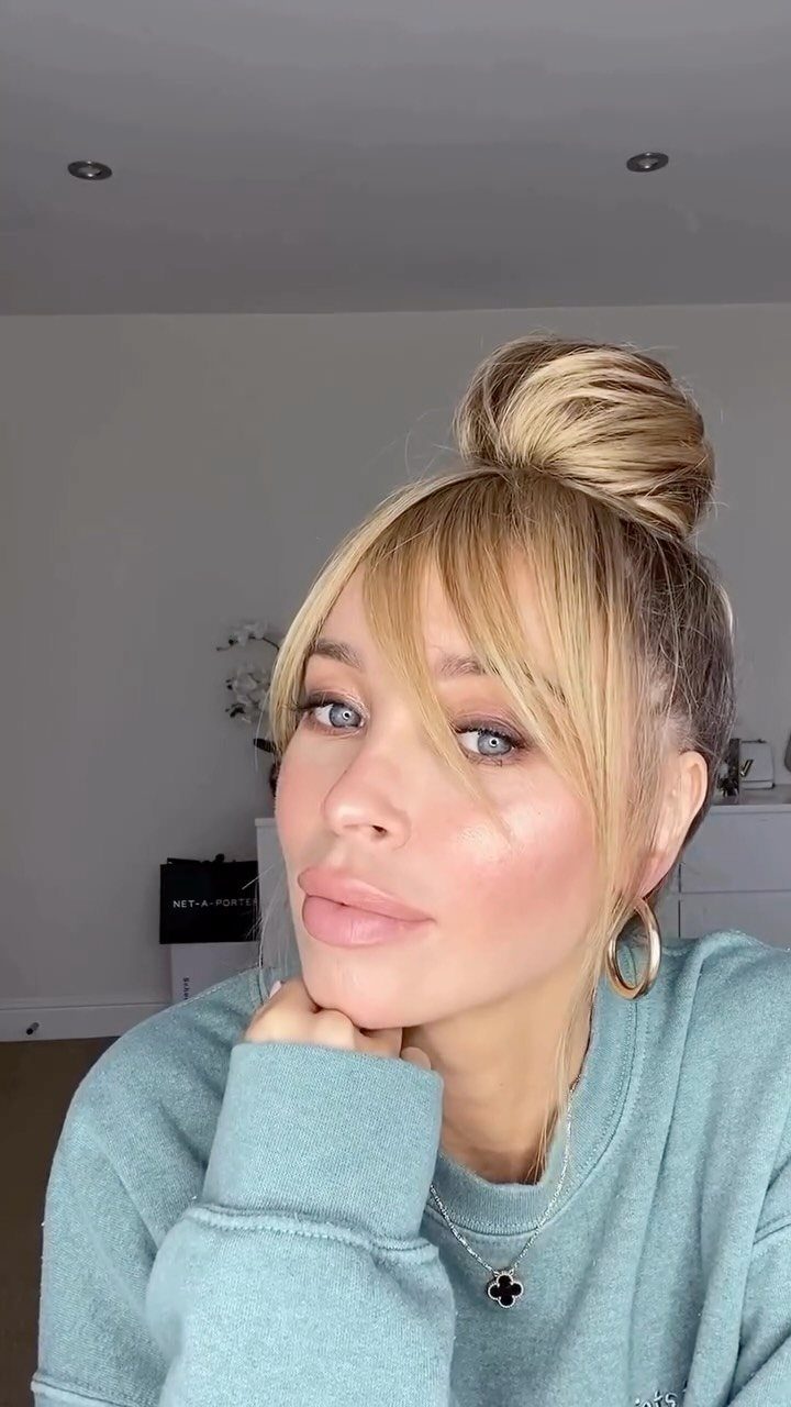 Woman with blonde hair styled in a high top knot bun and bangs.