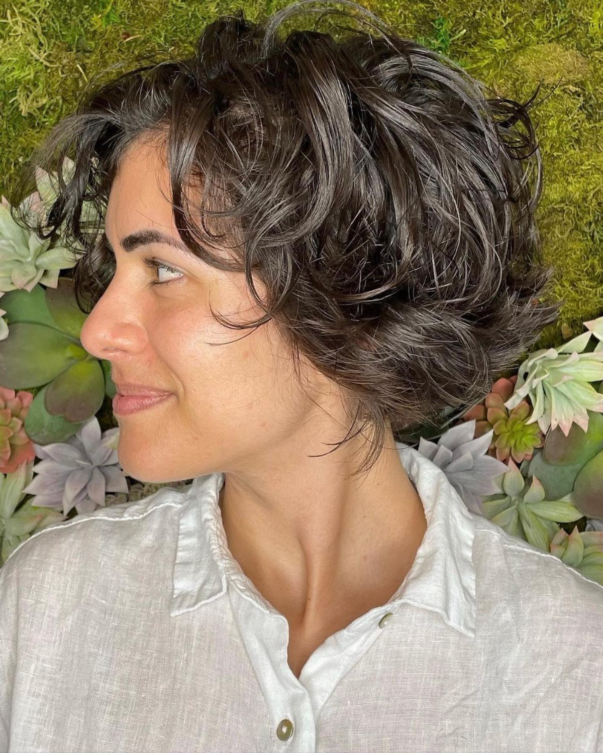 21 Short Curly Hair Styling Ideas for Every Texture 16 Woman with a short, curly bixie haircut posing against a succulent wall.