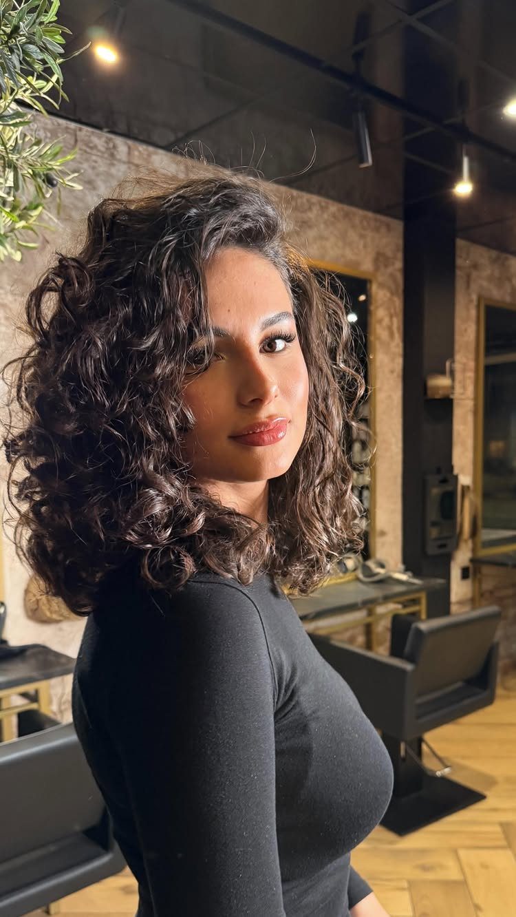 21 Short Curly Hair Styling Ideas for Every Texture 9 Woman with voluminous shoulder-length dark curly hair posing in a modern hair salon.