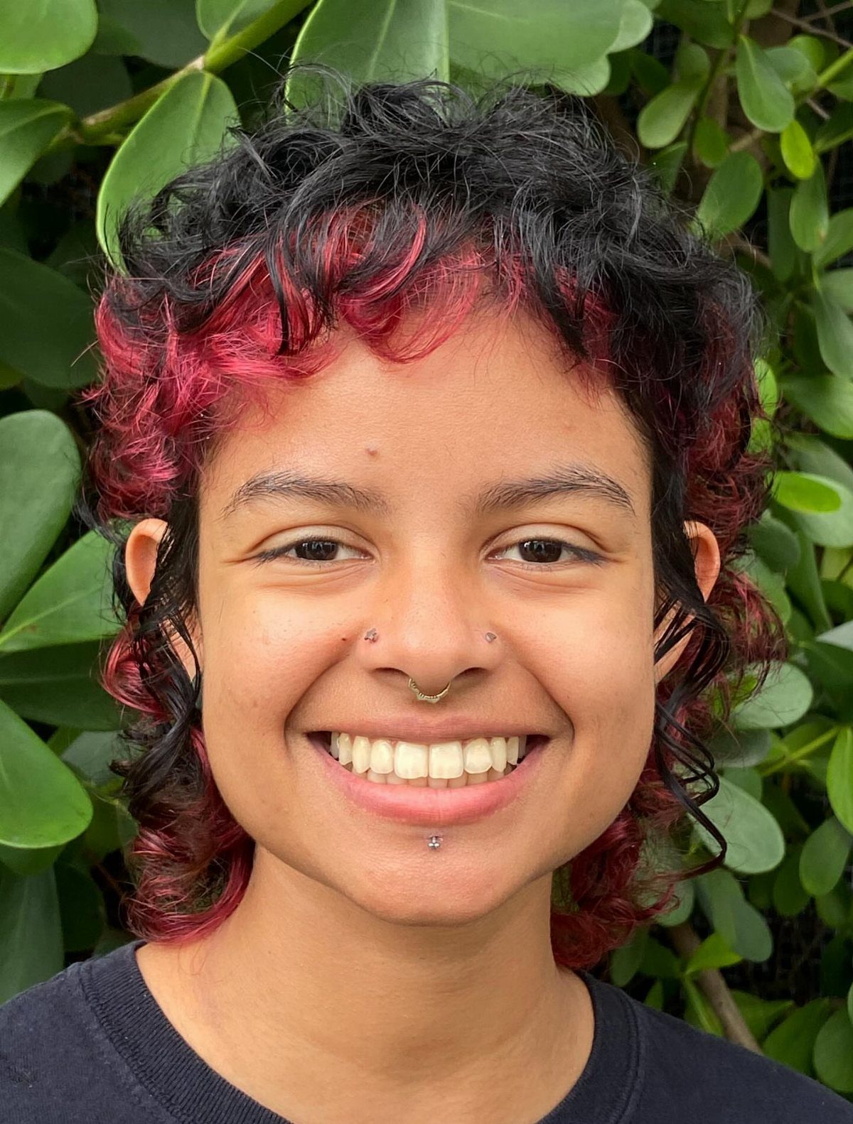 21 Short Curly Hair Styling Ideas for Every Texture 21 Person with short curly black and red hair smiling against green foliage