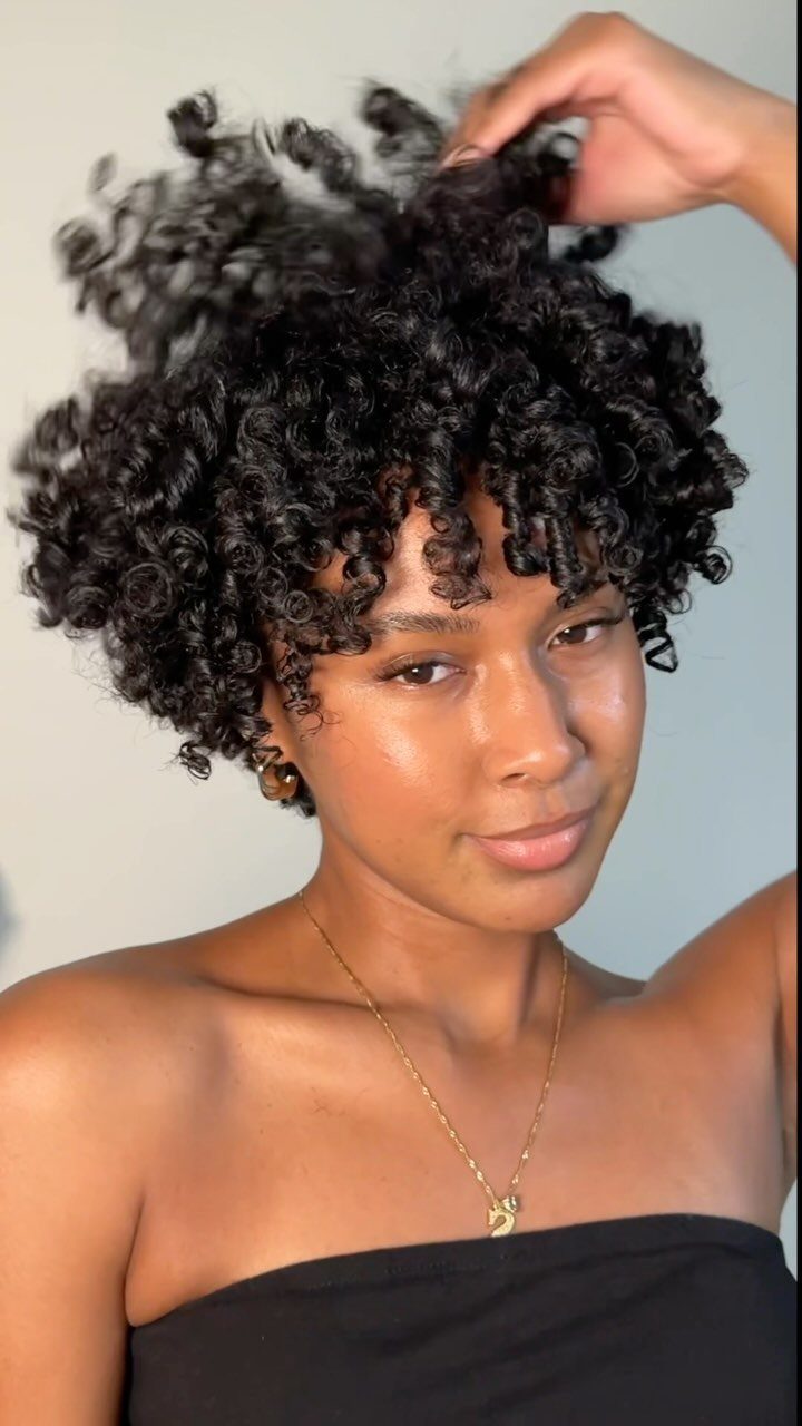 21 Short Curly Hair Styling Ideas for Every Texture 13 Woman with short voluminous black curly hair posing against a light grey wall.