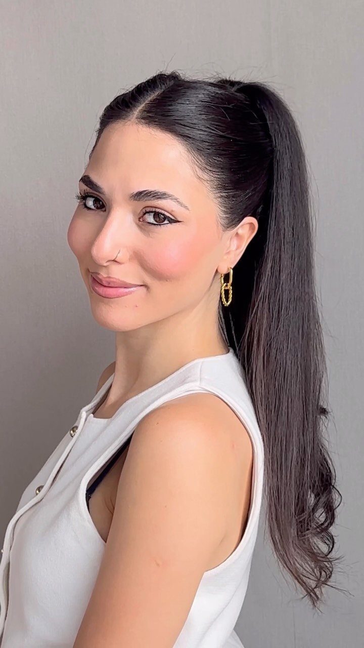 5 Sleek High Pony Ideas with Braids, Curls and Edges 1 Woman with dark hair styled in a high ponytail wearing a white top.