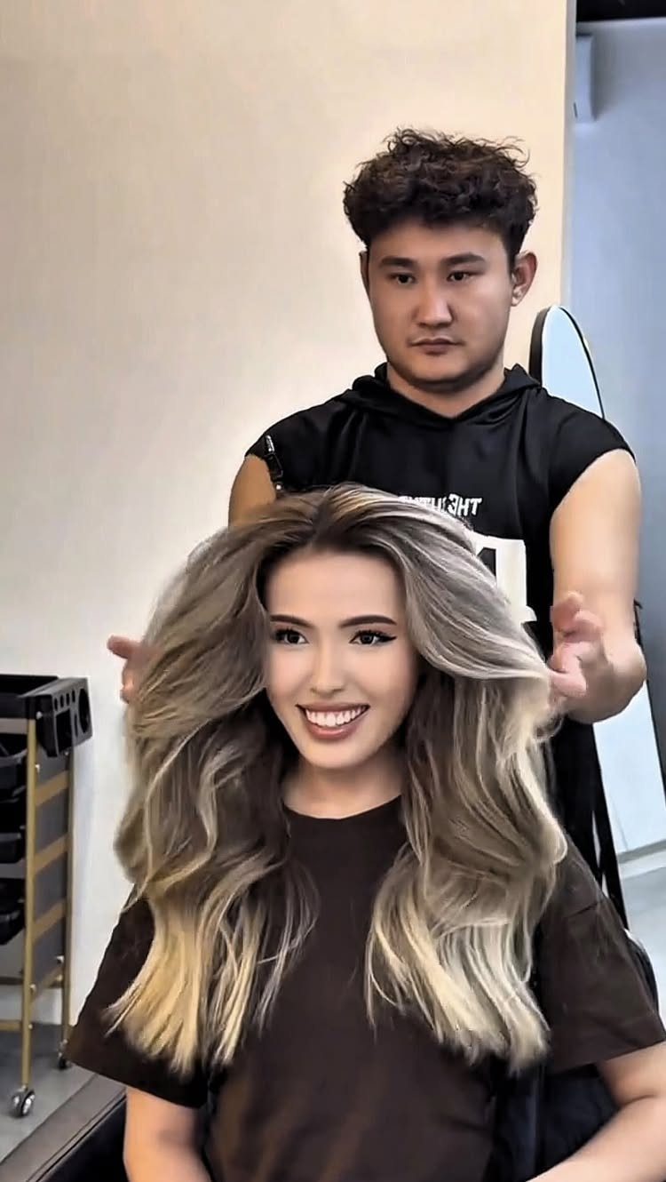 9 Toffee Caramel Hair Ideas for Brown and Blonde Bases 5 Hairstylist showcasing a woman's long wavy toffee caramel balayage hair in a salon.