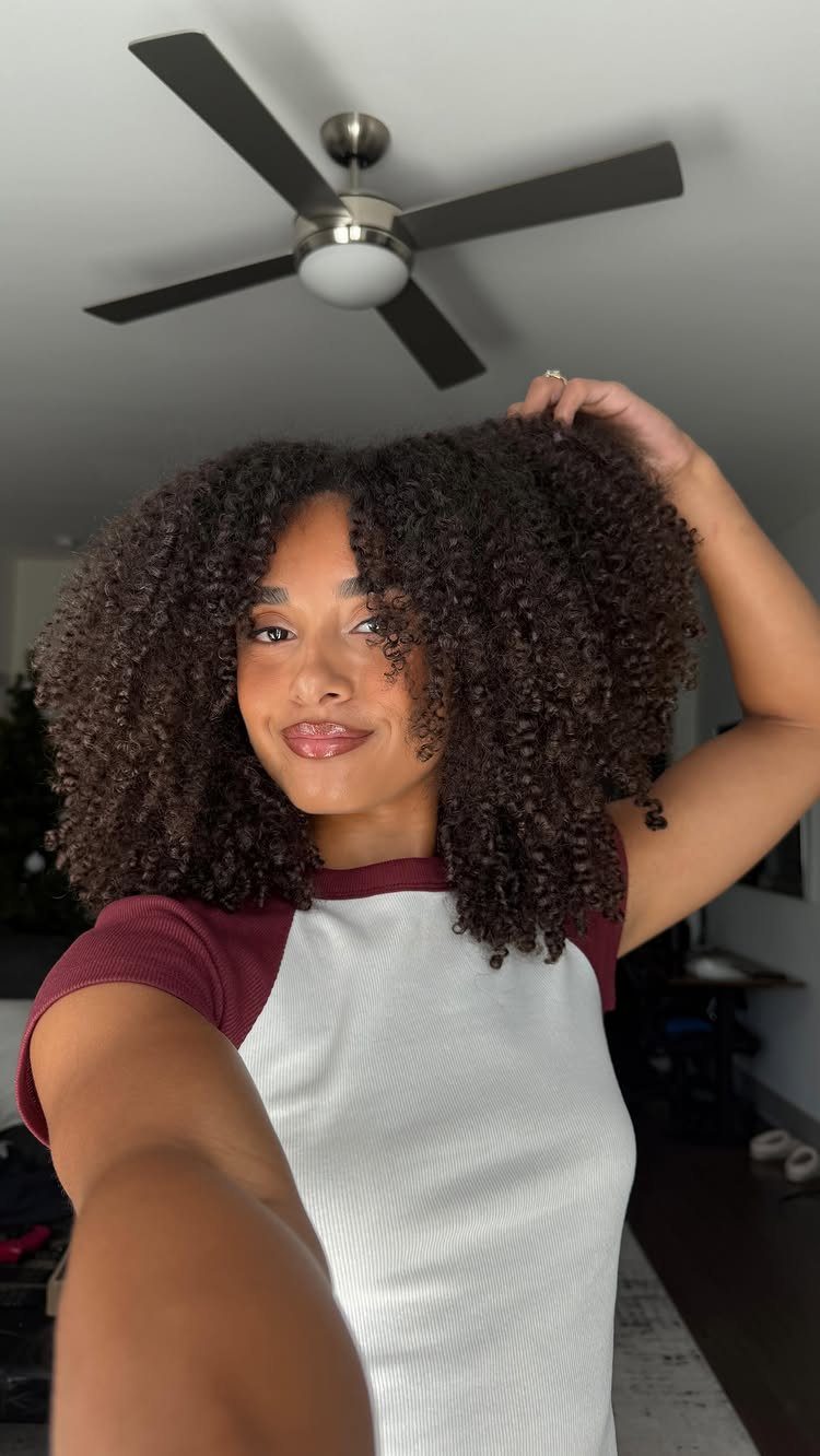 13 Trending Hair Style Ideas, from Curtain Bangs to Pixies 2 Woman with voluminous dark curly hair wearing a white and maroon raglan shirt.